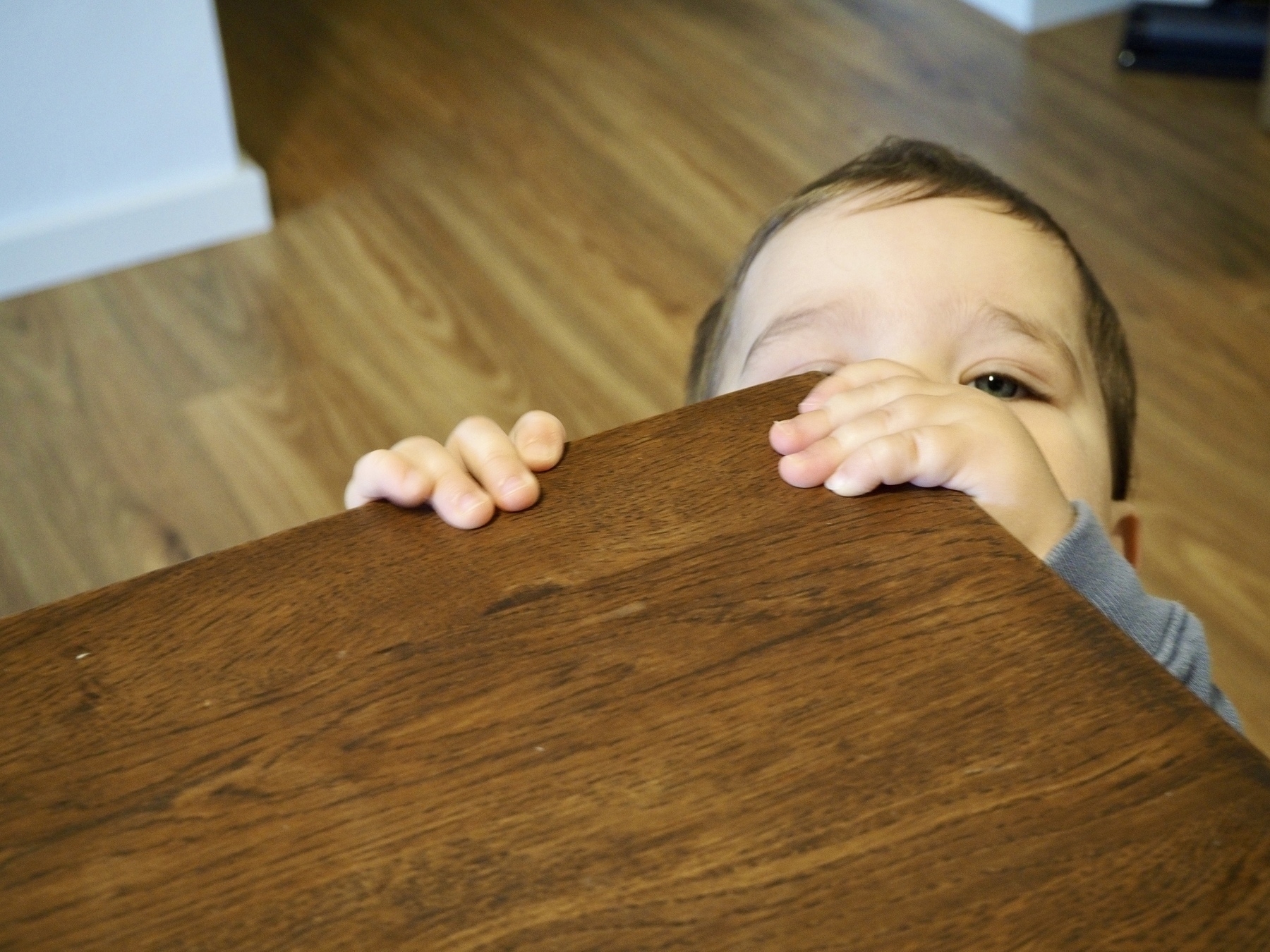 A toddler peers over the corner edge of a wooden dining table.