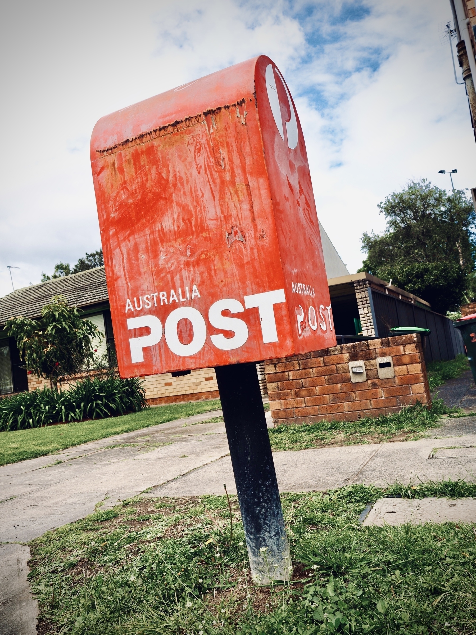 A rusty Australia Post box on a suburban footpath in front of a brick house