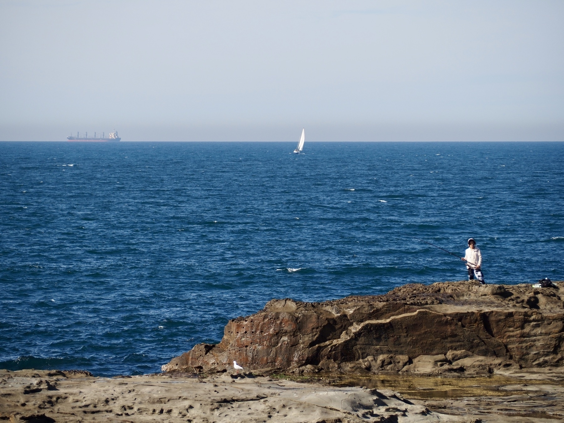 A person stands at the bottom-right of the frame on a rocky cliff, with a yacht and ship floating on the ocean in the distance.