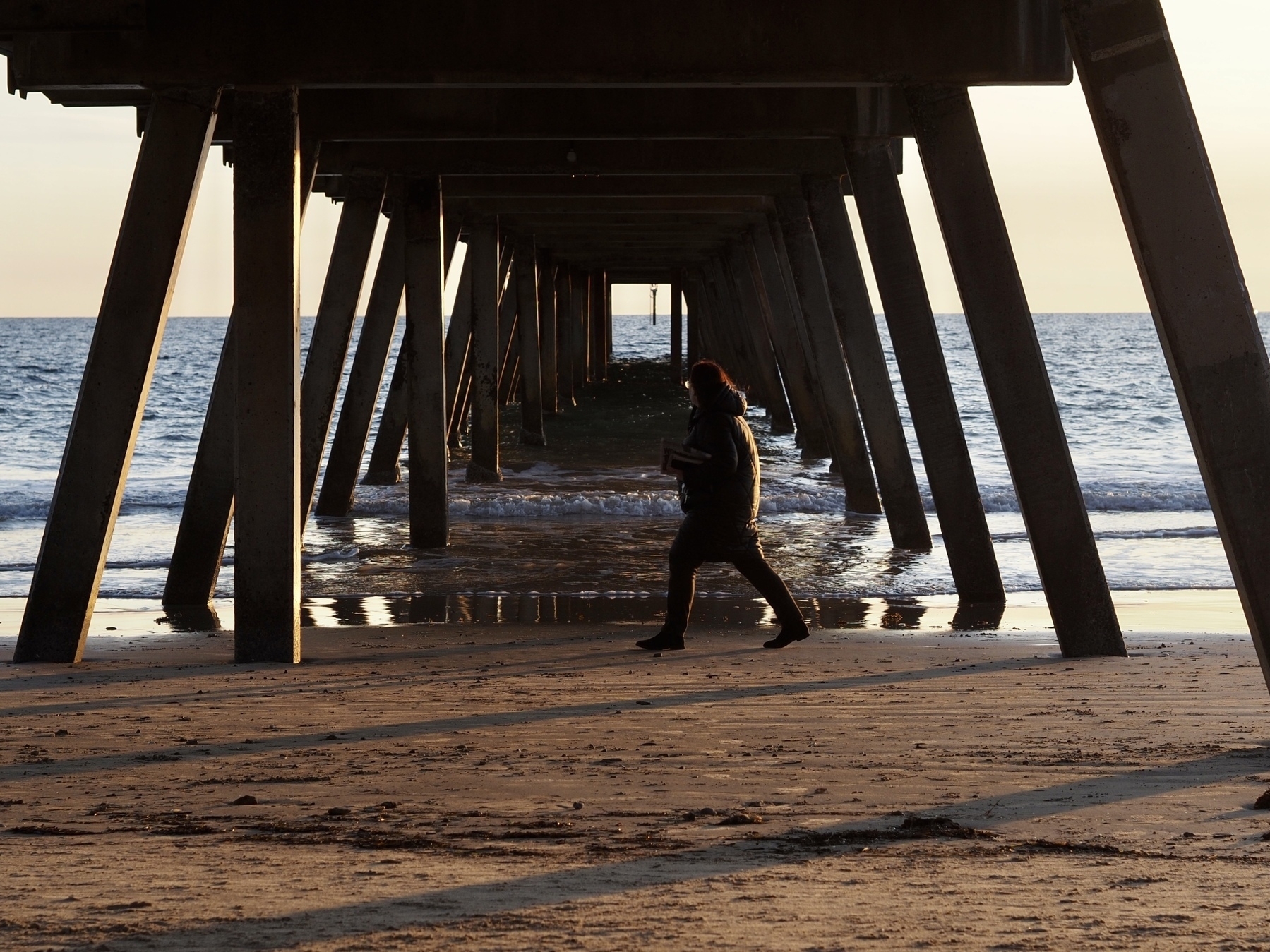 A woman walks under Glenelg Jetty, with water gently lapping on the sand.