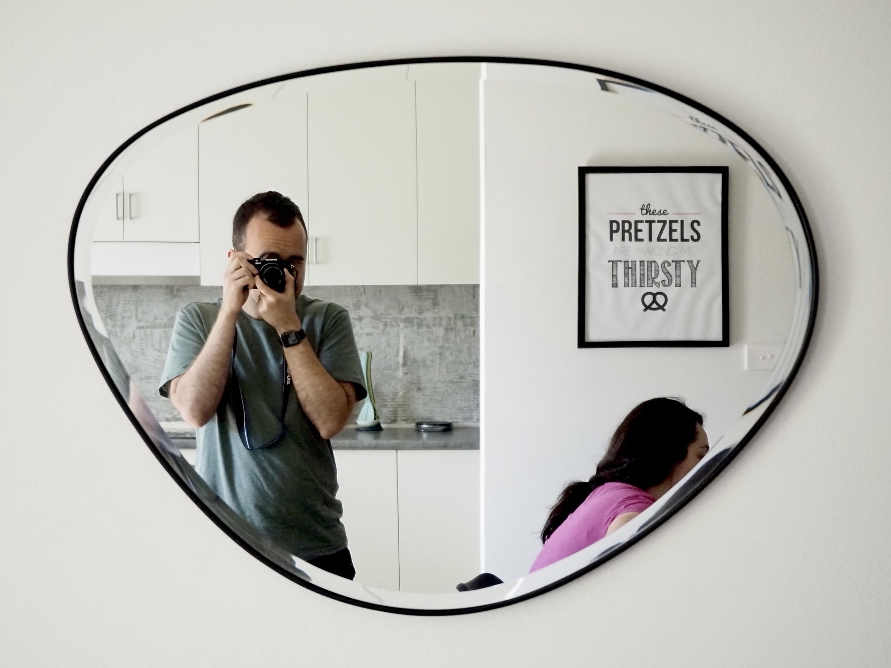 A man photographs himself and a woman in an oddly-shaped mirror with a kitchen bench in the background, along with a framed print that says 'These pretzels are making me thirsty'.